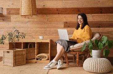 Young woman using laptop in armchair near wooden wall at home