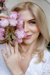 Naklejka premium Beautiful young blonde woman in a hat holding a sakura branch in front of her