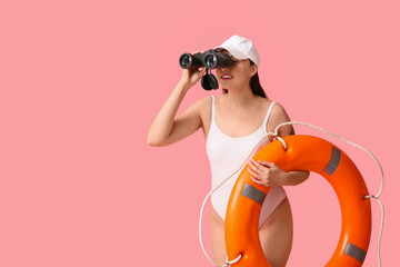 Young female lifeguard with ring buoy looking through binoculars on pink background