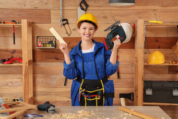 Young female carpenter with cordless jigsaw and wooden plank in workshop. Labor Day celebration