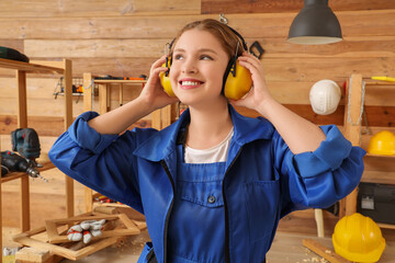 Young female carpenter with earmuffs in workshop. Labor Day celebration - obrazy, fototapety, plakaty