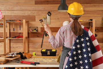 Young female carpenter with USA flag and wrench in workshop, back view. Labor Day celebration