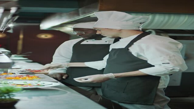 Vertical shot of Caucasian master chef in apron and gloves serving food on plate and teaching African American apprentice how to work in kitchen of fine dining restaurant