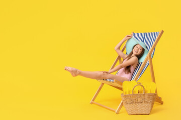 Cute little happy girl in swimsuit with wicker bag and flippers sitting on deckchair against yellow...