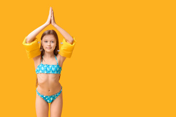 Cute little girl in swimsuit with inflatable armbands on yellow background
