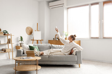 Young woman turning on conditioner with remote control and lying on sofa in living room