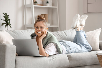 Young woman in eyeglasses lying on sofa and chatting with laptop in living room