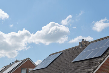 Blue solar cells or sun panels on roofs of houses in a row reflecting the blue cloudy sky. Green...
