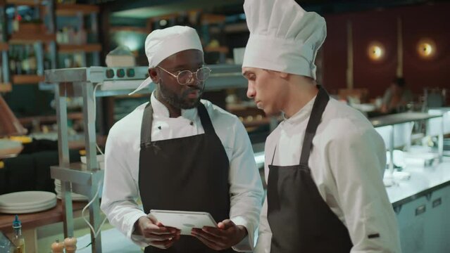 Head chef cooking food and then giving job task to colleague using digital tablet while working together in restaurant kitchen