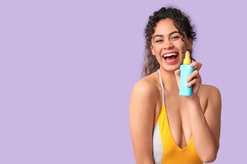 Happy young African-American woman in swimsuit with bottle of sunscreen cream on lilac background