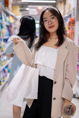 A smiling Asian woman stands in a grocery aisle, holding shopping bags, shopping in a supermarket.