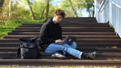 Young man sitting on the staircase in the city park while using laptop and typing. Autumn season. Education, working concept. Slow motion