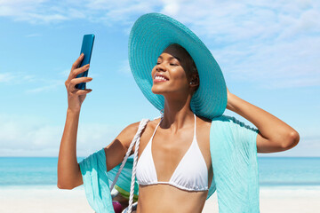 Happy woman at the beach side wearing bikini, blue sun hat and pareo and using mobile phone in a...