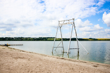 wooden swing and bridge in the water on a summer lake, beach, sand, sunbeds, white clouds