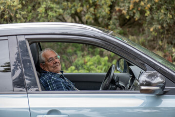 older man sitting in the driver's seat waiting, light blue 4x4 car in the bush. Person dressed in blue shirt, grey hair and glasses.