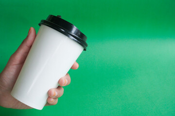 A hand holds an white disposable cup of tea or coffee on a green background. Blank space for...