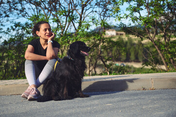 Multi ethnic beautiful pensive woman sitting next to her dog on the asphalt, a beautiful black...