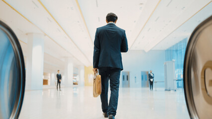 Fototapeta premium Back view of skilled business man phone calling to project manager while walking to meeting room. Caucasian project manager walking with meeting room with blurred background. Full body. Exultant.