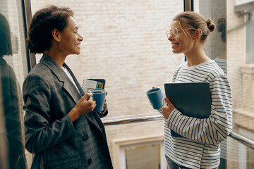 Two smiling female colleagues have a coffee break time while standing at office terrace