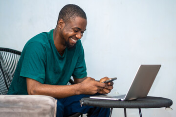 photo of young African American guy working on laptop and using smartphone. African man wearing green shirt, Texting, Internet, Technology. Mobile phone © WISDOM