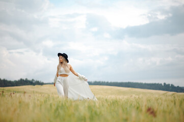 Stylish blonde woman in white dress and black hat posing on the summer green field. Outdoor photoshoot