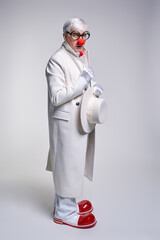 Serious, suspiciously looking clown in a white coat and large red patent leather clown boots with...