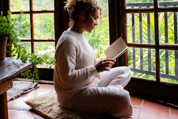 Serene woman reading a book at home sitting on the floor in total mindful chilling indoor leisure...