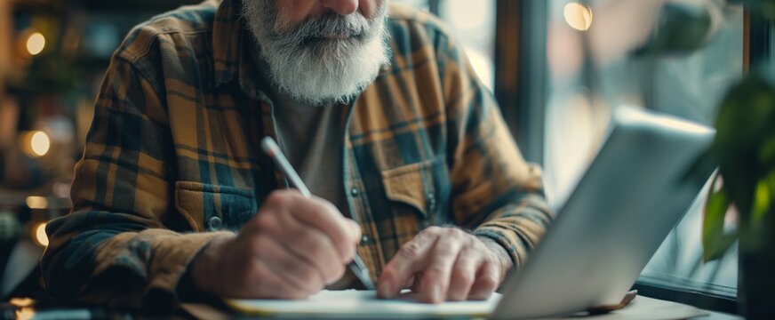 Elderly bearded man in plaid shirt pensively writing in a notebook with a laptop, representing creativity or lifelong learning