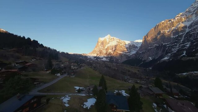 Rugged cliff face illuminated by sunset light. Drone flying forward over deep shaded valley in Swiss Alps. Grindelwald landscape aerial view.