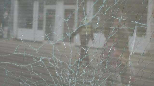 50fps. CU Broken, shattered shop window front. Violence, crime, vandalism. People and police officer reflected in the broken glass window. 