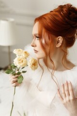 Naklejka premium Elegant lady with bright red hair, clad in a white dress, posing gracefully with a bouquet of white roses indoors