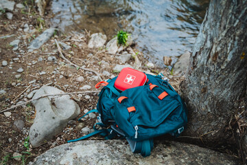An electric blue backpack with a red first aid kit is by the river on a rock