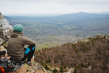 Man sits on mountain edge, gazing at valley below under cloudy sky