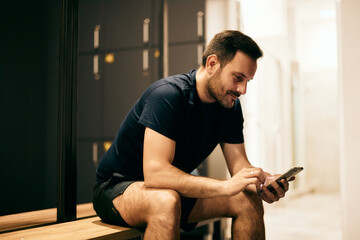 The fitness man finished his workout, sitting in the locker room, using a mobile phone and smiling.