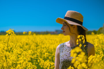 Brunette girl in straw hat, enjoying the view of blooming rapeseed field in background, embracing...