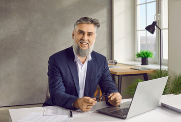 Portrait of happy bearded mature business man in suit working at the desk on his workplace with a...