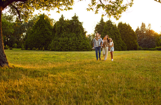 Happy family spending time outdoors. Parents and kids strolling in nature. Mother, father and children enjoying quiet summer evening and walking together on green lawn in beautiful city park or garden - Powered by Adobe