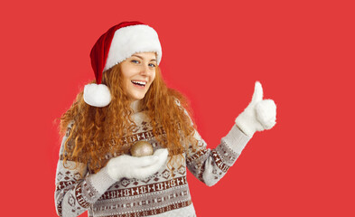 Naklejka premium Girl is advertising Christmas sale or special offer. Portrait of cheerful cute young woman holding Christmas tree toy in her hand and showing thumb up. Woman in Santa hat isolated on red background.