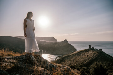 A woman stands on a rocky hill overlooking the ocean. She is wearing a white dress and she is enjoying the view. The scene is serene and peaceful, with the sun shining brightly in the background.