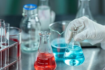 A gloved hand holding a blue liquid-filled flask over a table with other lab equipment.