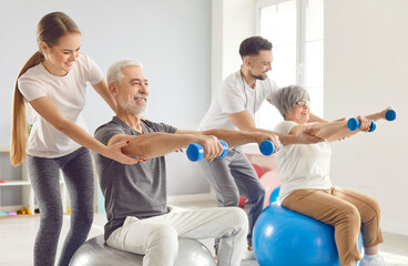 Coach or health care worker helping mature people to do sport exercises with dumbbells sitting on fit ball in gym. Physiotherapist helping retired man and woman in rehab in rehabilitation center.