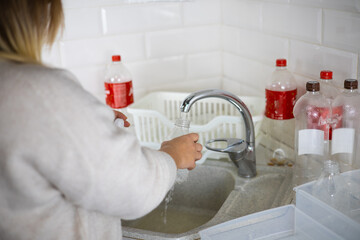 A woman standing with her back is washing a plastic bottle in the sink A woman sorts plastic in the...