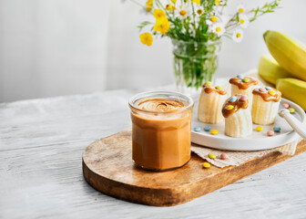 Slice banana pieces with peanut butter, colorful sprinkles, and a knife on a light wooden table. Empty space for text. Creative idea for kid breakfast, healthy snack for kids.