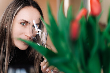 Young woman looks mysteriously at camera against the background of a blurred bouquet of flowers, covering one eye with a wine glass