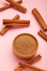 Cinnamon sticks and powder in wooden bowl on pink background.