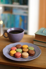 Purple plate filled with pastel macarons, cup of tea or coffee, vintage books and reading glasses on the table. Colorful bookcase in the background. Selective focus.