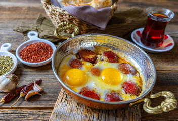 Turkish sausage fried in a copper pan, egg, served with tea and bread, wooden background, close-up.