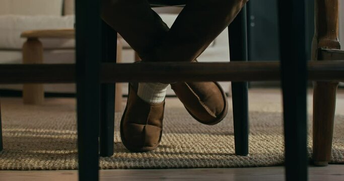 A man is sitting at home at a table. Close-up of his feet in slippers. An unrecognizable person