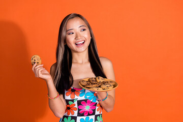 Photo of toothy beaming asian woman wear print dress holding cookie look at discount empty space isolated on orange color background