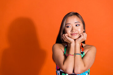 Photo of cute woman with straight hair wear print dress look at promotion empty space arms on cheeks isolated on orange color background